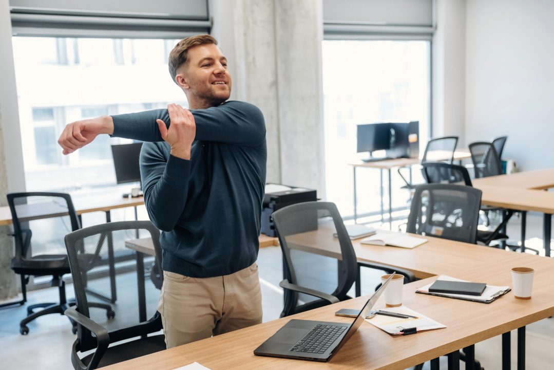 Young man in a blue sweater stretches his arm in a modern office setting, featuring wooden desks, laptops, and ergonomic chairs in the background